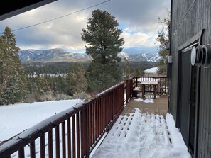 Un muelle de madera con barandilla cubierta de nieve, un banco y vistas a montañas y árboles nevados.