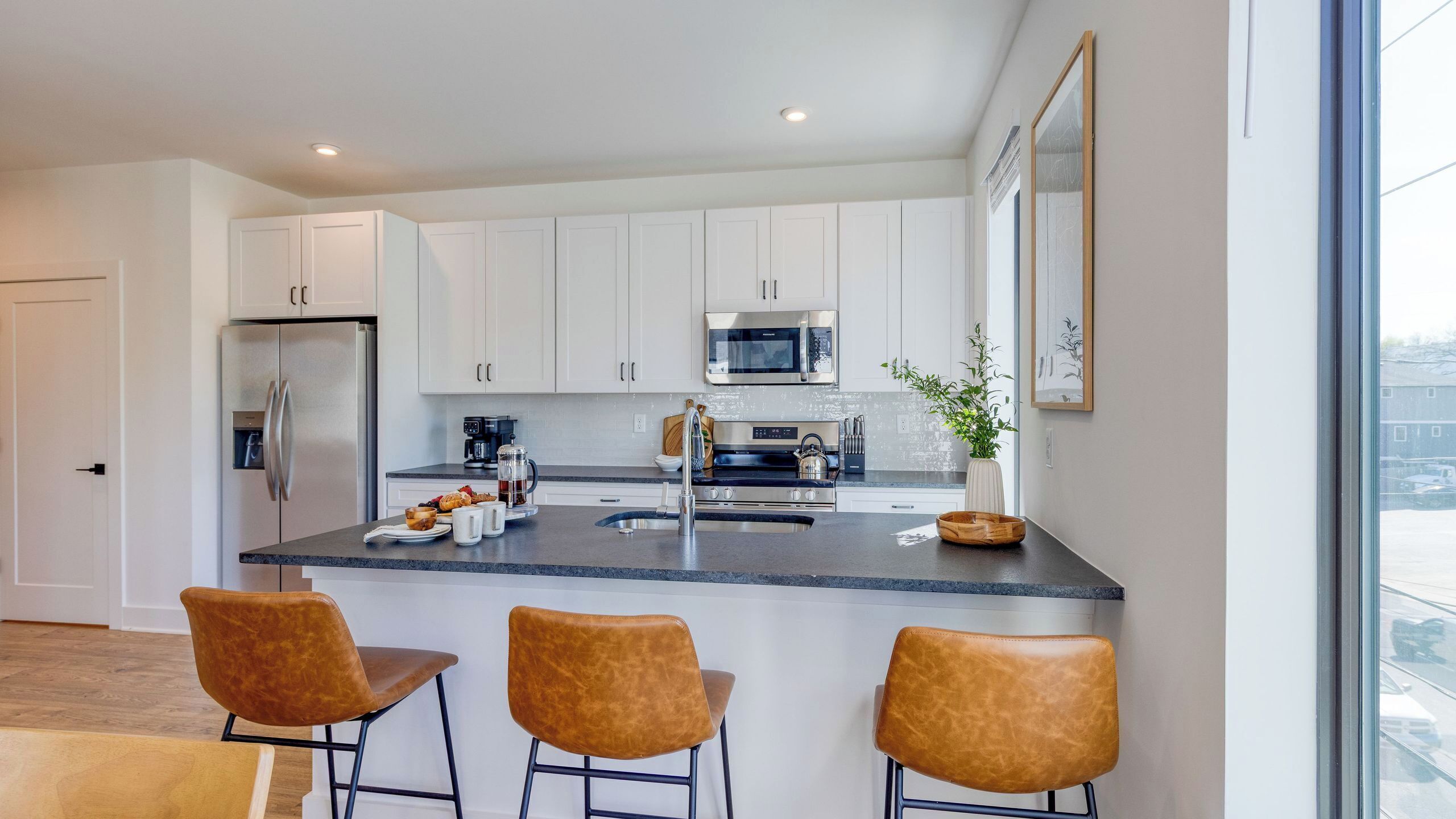 A modern kitchen with white cabinets, stainless steel appliances, a central island with bar stools, and a large window.