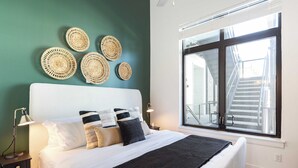 A neatly made bed with a black throw blanket, white bedding, and multiple pillows. There are woven wall decorations above the bed and a large window with a view of a staircase. - The Heights by AvantStay (Nashville)