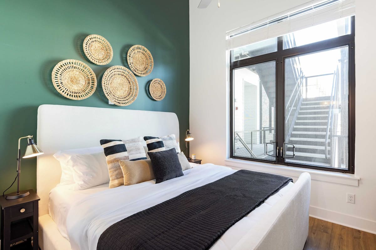 Una cama bien hecha con un edredón negro, ropa de cama blanca y varias almohadas. Hay decoraciones de pared tejidas sobre la cama y una ventana grande con vista a una escalera.