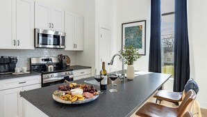 A modern kitchen with white cabinets, stainless steel appliances, a large island with a fruit and cheese platter, wine glasses, and a potted plant. - The Heights by AvantStay (Nashville)