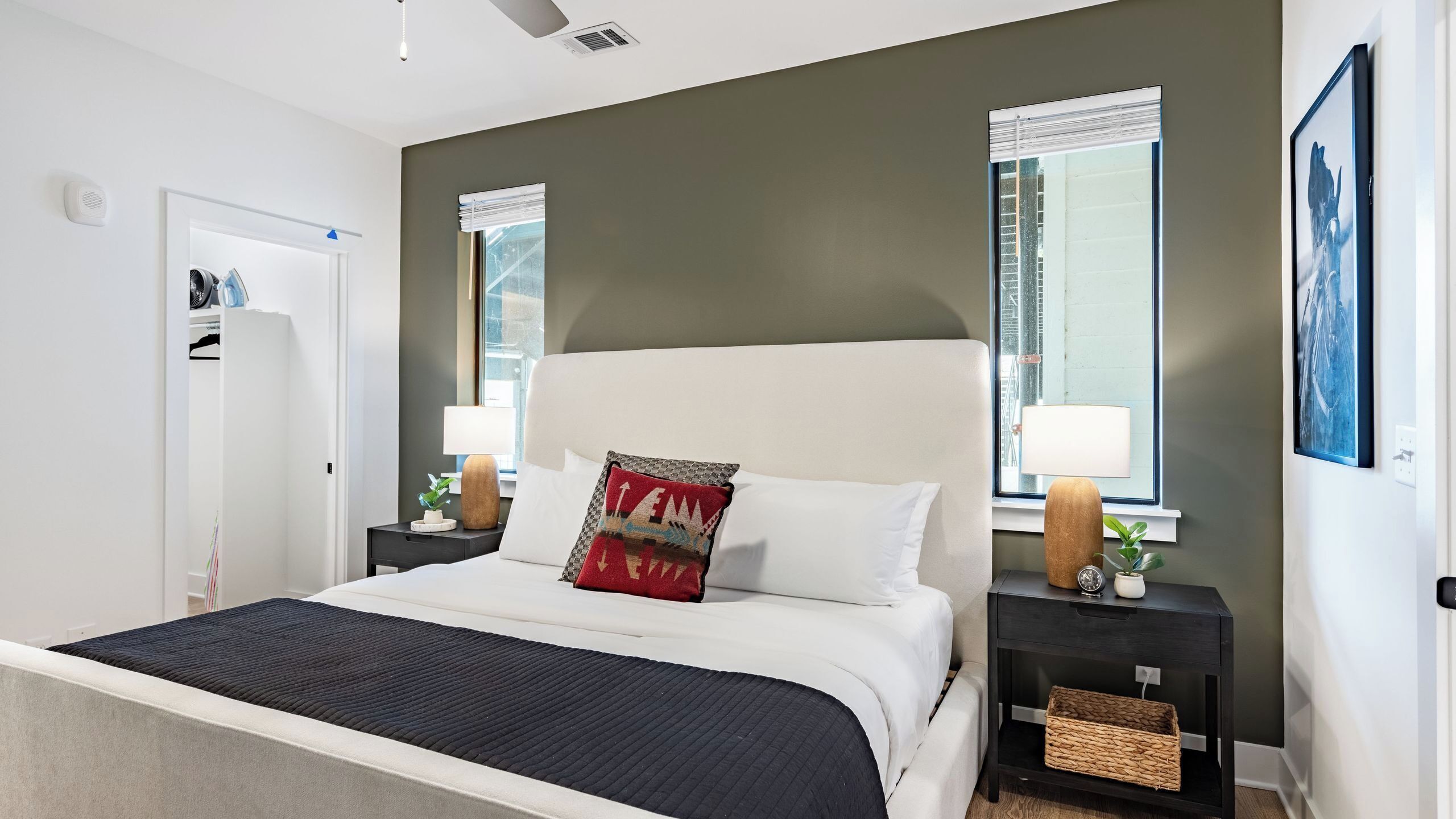 A modern hotel room with a large bed, a nightstand with a lamp, a small side table, and a window with blinds.