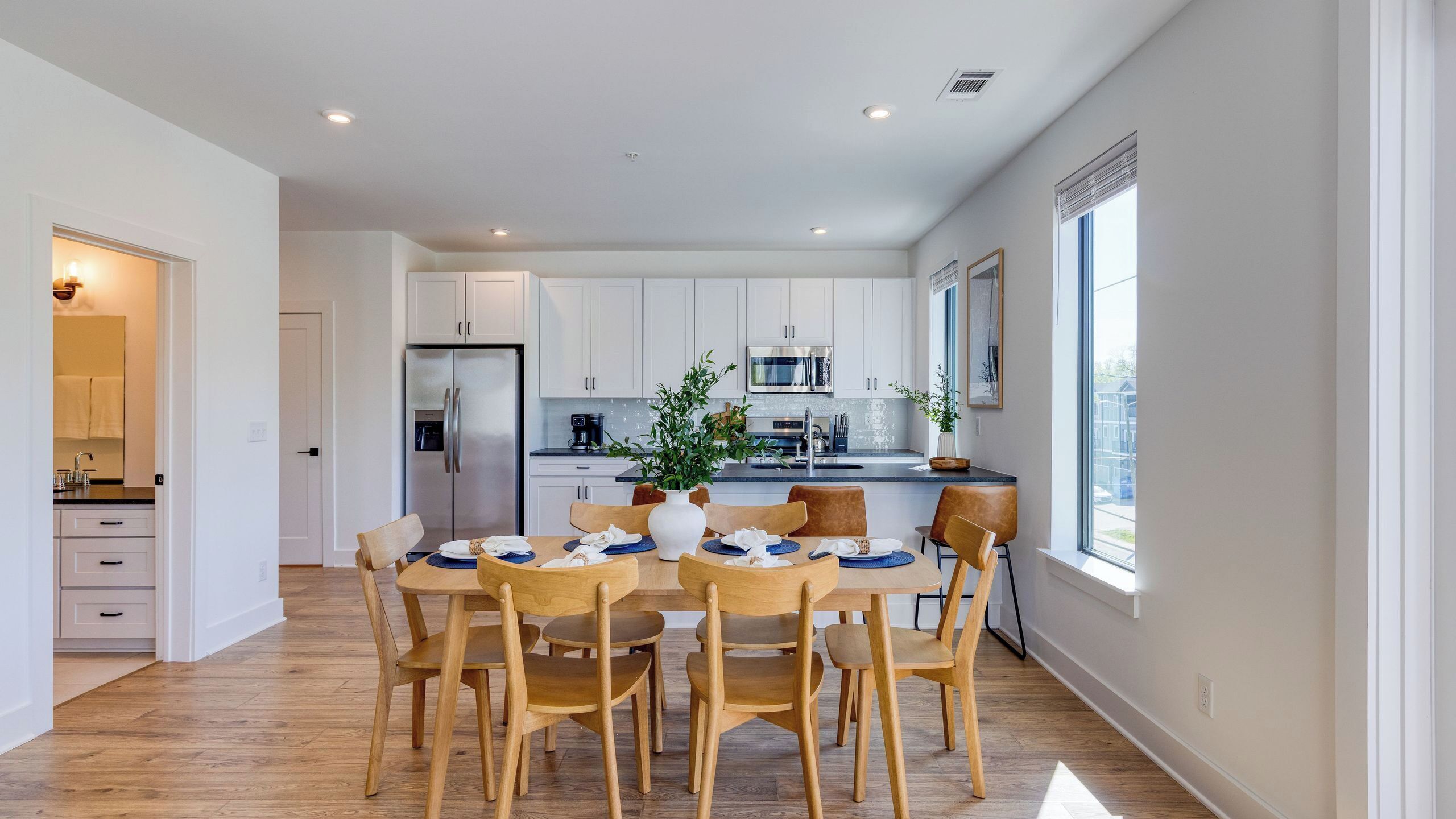 Una cocina moderna con armarios blancos, un refrigerador de acero inoxidable y una zona de comedor con sillas y mesa de madera.