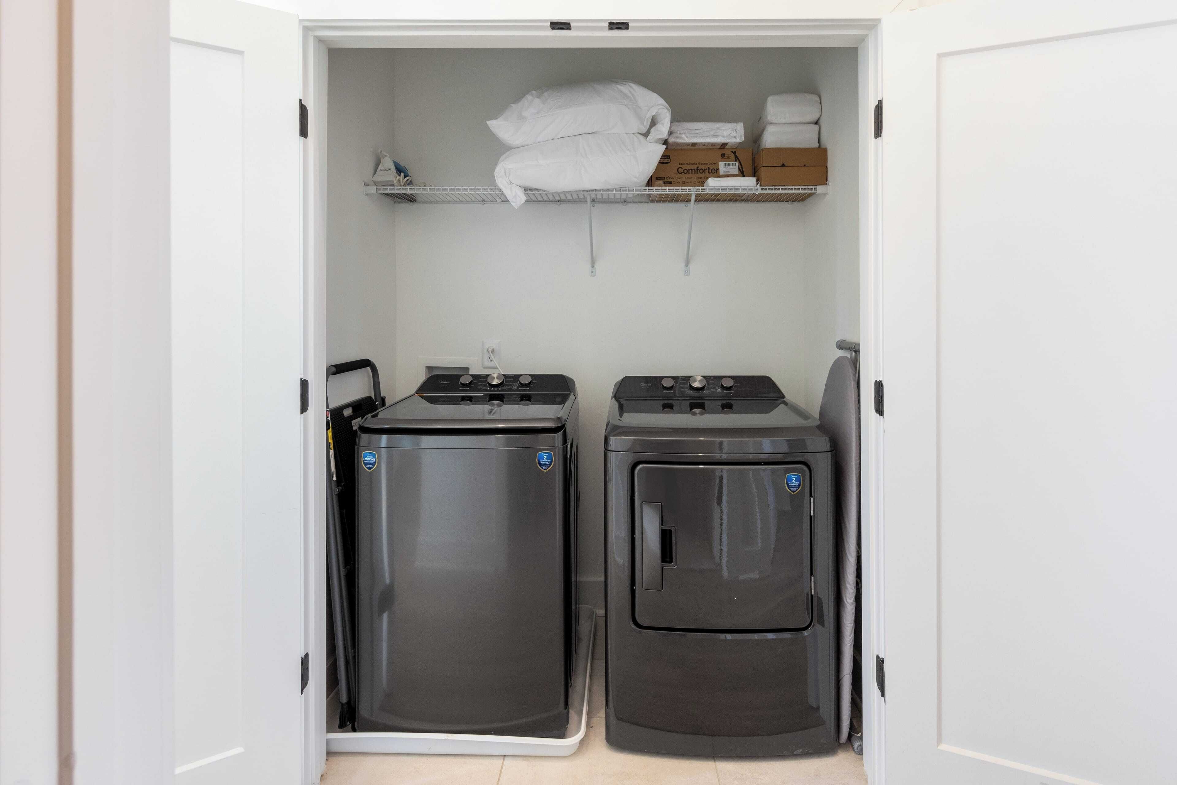 A compact laundry room with two wall-mounted washing machines and a shelf above containing folded towels and a box.