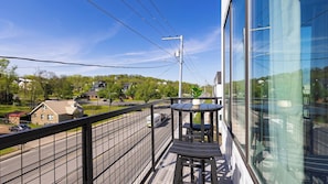 Un balcon avec une table et des chaises, donnant sur une rue animée où l’on peut voir des voitures et des maisons, et un ciel bleu dégagé.