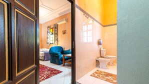 A hotel room with a wooden door, a blue armchair, a bed with patterned bedding, a window with decorative curtains, and a bathroom with a toilet and sink. - Riad Qodwa (Sidi Abdallah Ghiat)