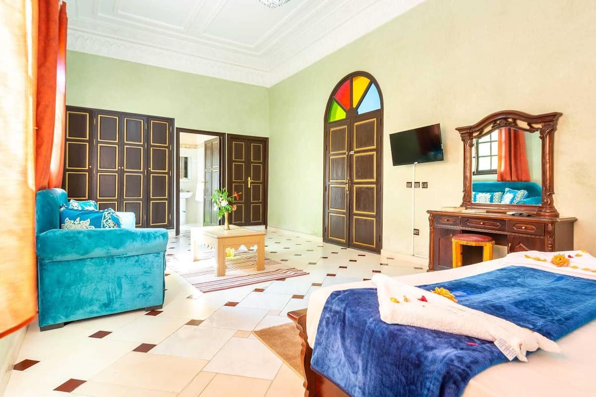 Quarto de hotel com uma cama grande, um sofá azul, uma cômoda de madeira, uma TV e um vitral colorido.