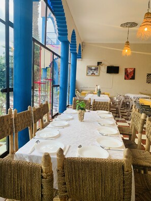 A dining area with tables set for a meal, blue pillars, wicker chairs, and a view through large windows. - Riad Qodwa (Sidi Abdallah Ghiat)