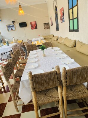 A dining area with a long table set for a meal, surrounded by wicker chairs, with a sofa and a television in the background. - Riad Qodwa (Sidi Abdallah Ghiat)