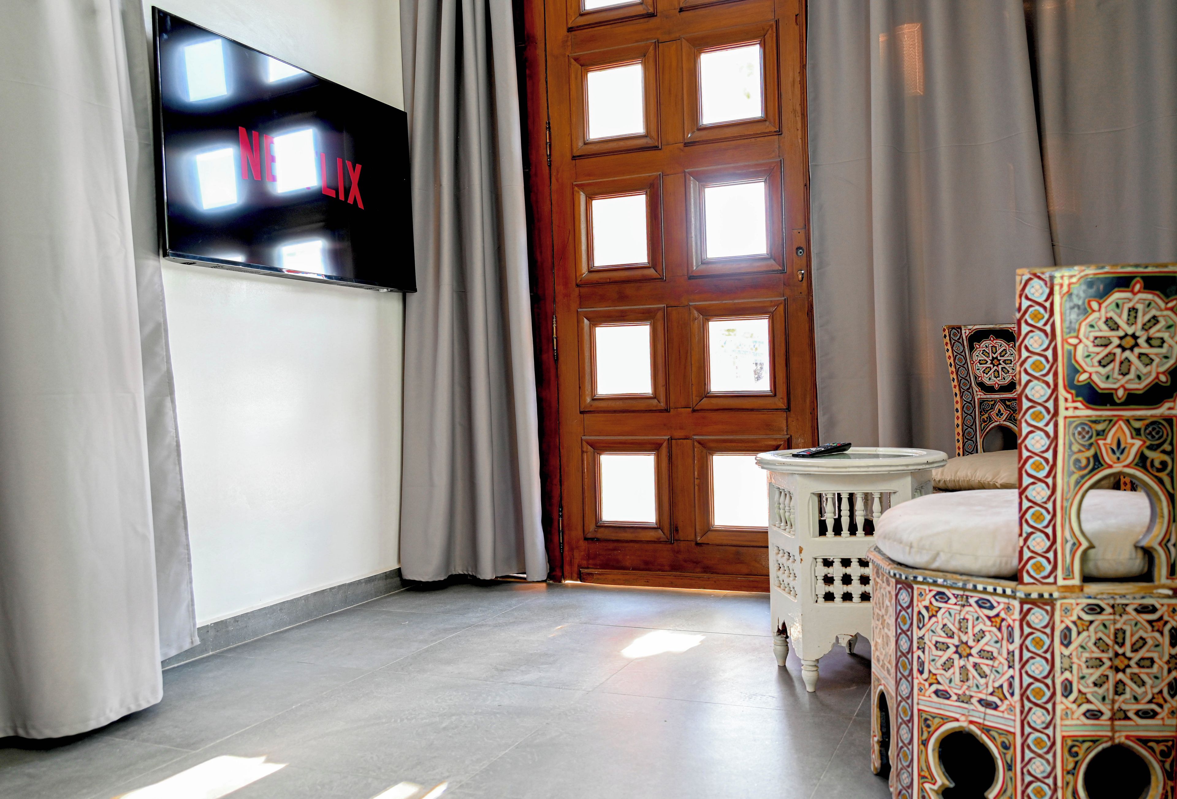 A room with a flat-screen TV mounted on the wall, a wooden door with multiple windows, and a patterned ottoman.