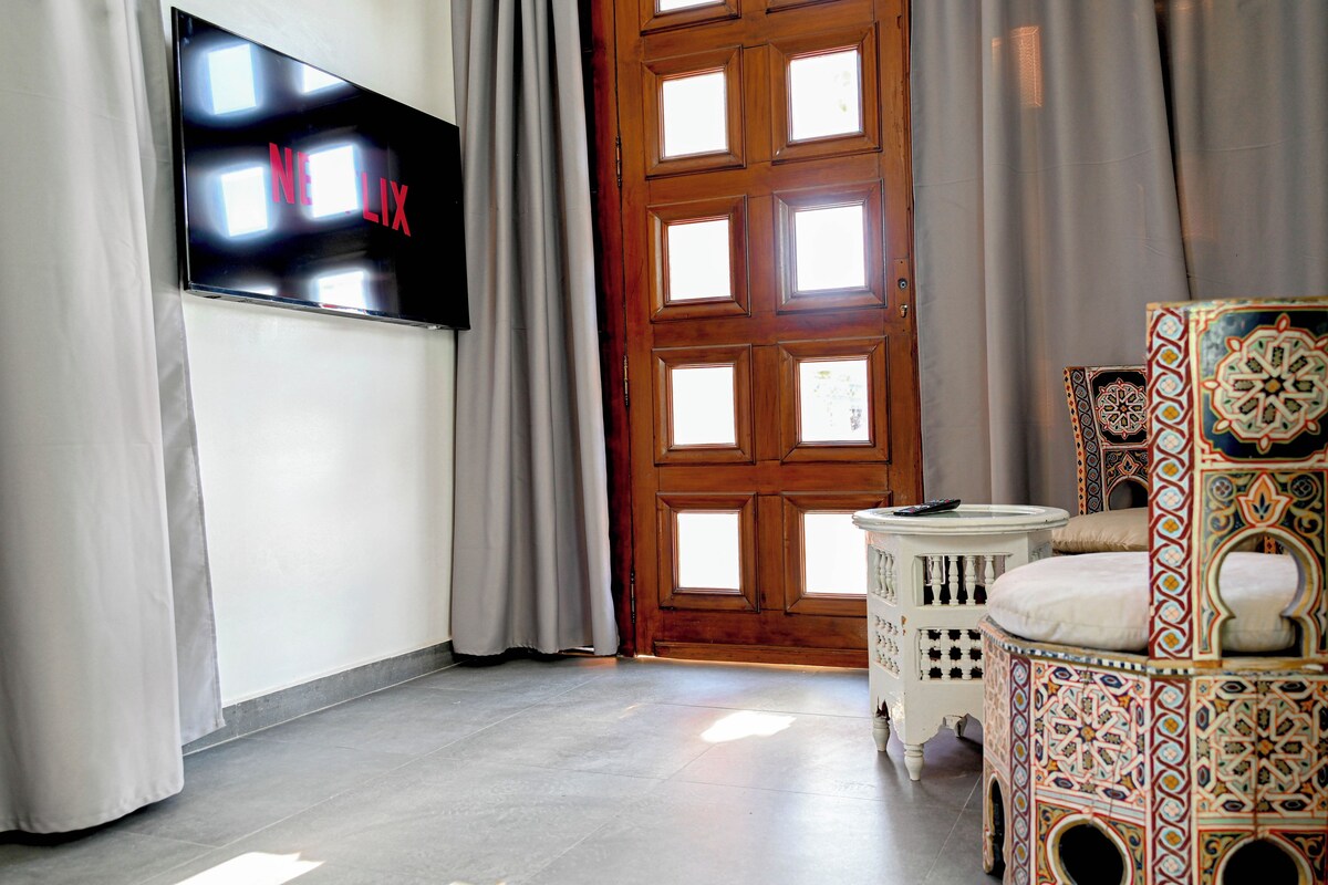 A room with a flat-screen TV mounted on the wall, a wooden door with multiple windows, and a patterned ottoman.