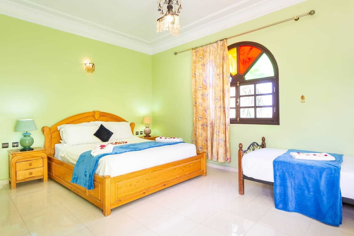Quarto com uma cama grande, uma cama menor, uma mesa de cabeceira com abajur e uma janela com cortinas.
