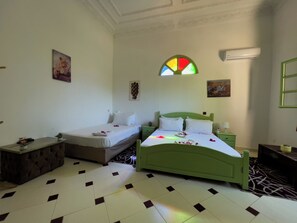 A hotel room with two single beds, a green headboard, a small bedside table, a nightstand, a wall-mounted air conditioner, and a decorative stained glass window. - Riad Qodwa (Sidi Abdallah Ghiat)