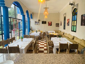 Un ristorante con soffitti alti, pilastri blu e pavimento a scacchi. I tavoli sono apparecchiati con tovaglie bianche e sedie.