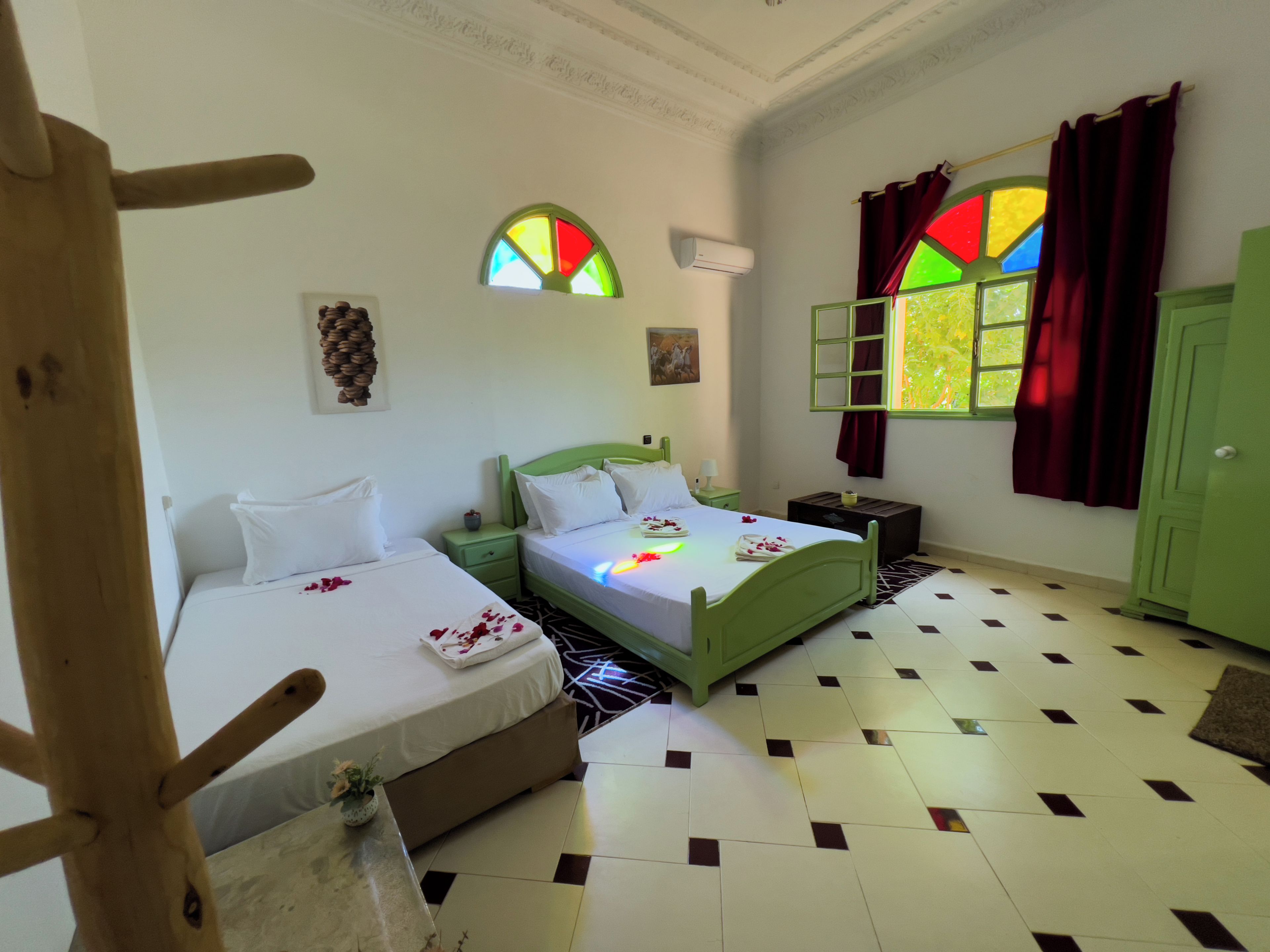 A hotel room with two beds, a green wardrobe, a window with colorful stained glass, and a patterned floor.
