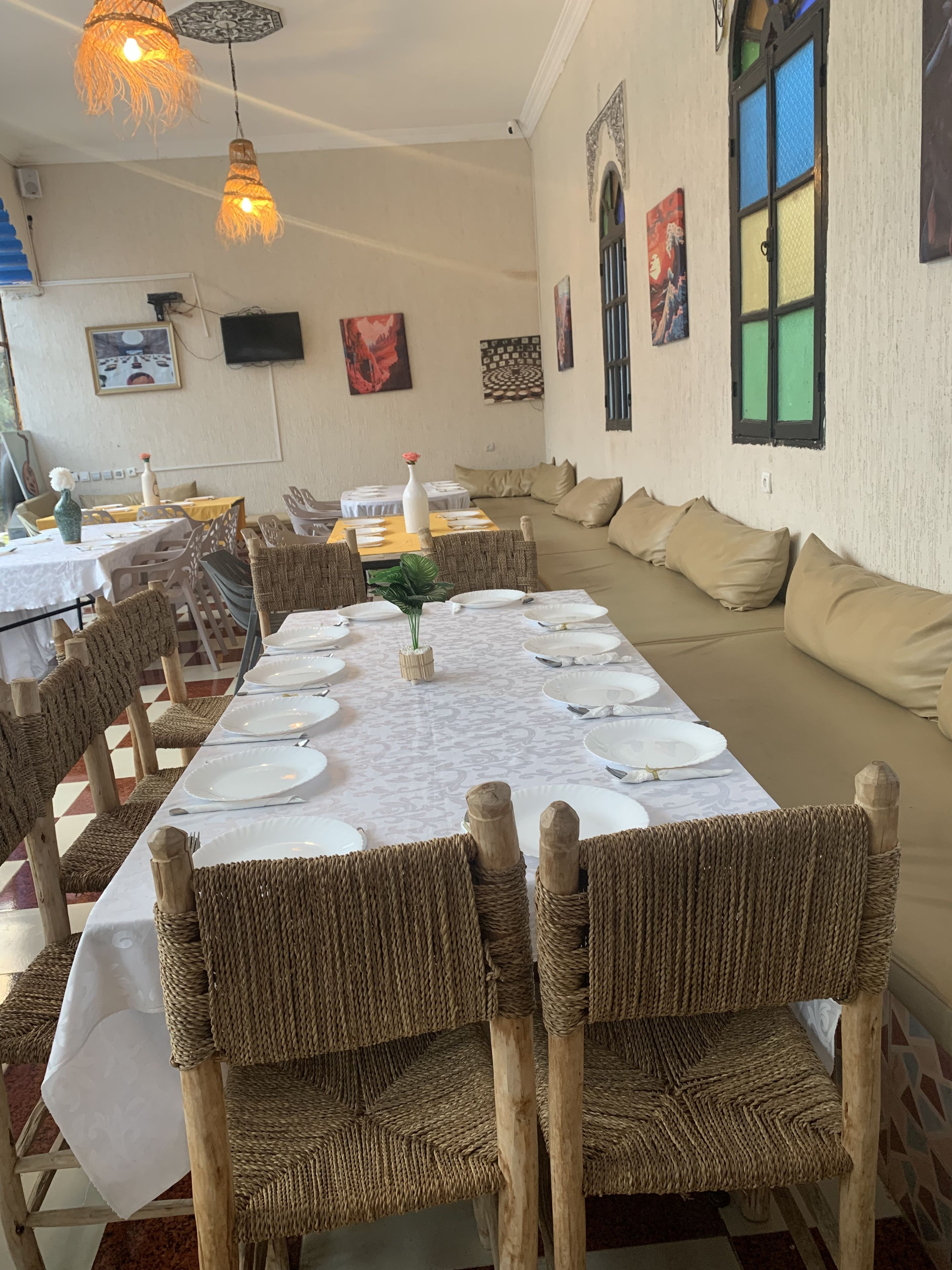 Uma sala de jantar com uma mesa comprida posta para uma refeição, rodeada por cadeiras de vime. Há luminárias pendentes sobre a mesa e quadros nas paredes.