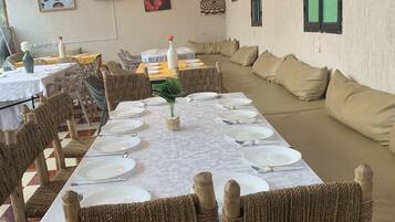 Uma sala de jantar com uma mesa comprida posta para uma refeição, rodeada por cadeiras de vime. Há luminárias pendentes sobre a mesa e quadros nas paredes.