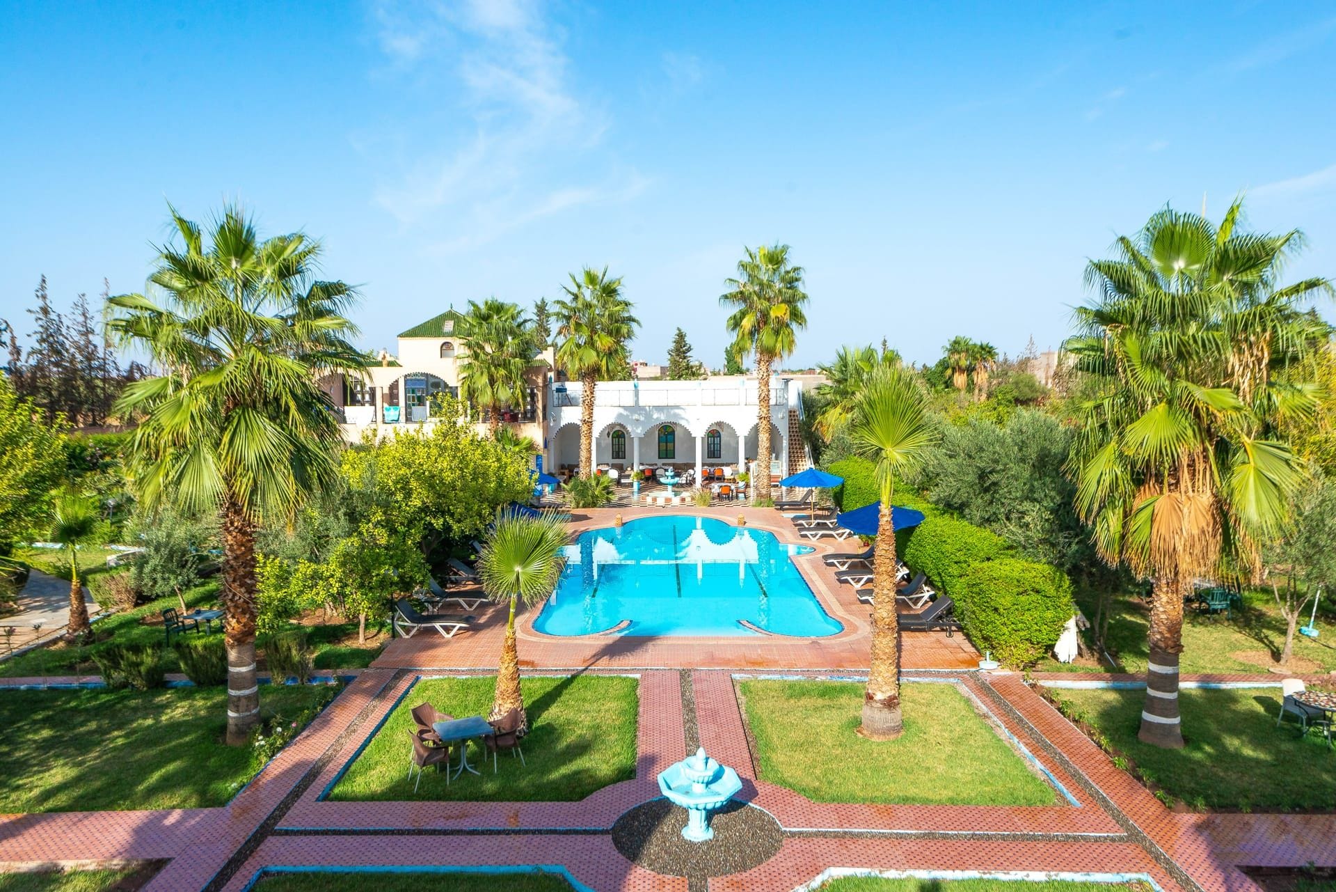 Zona de piscina con sillas de descanso y sombrillas, rodeada de palmeras y un edificio con arcos.