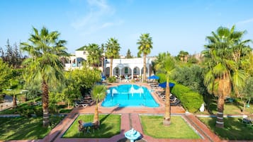 Zona de piscina con sillas de descanso y sombrillas, rodeada de palmeras y un edificio con arcos.