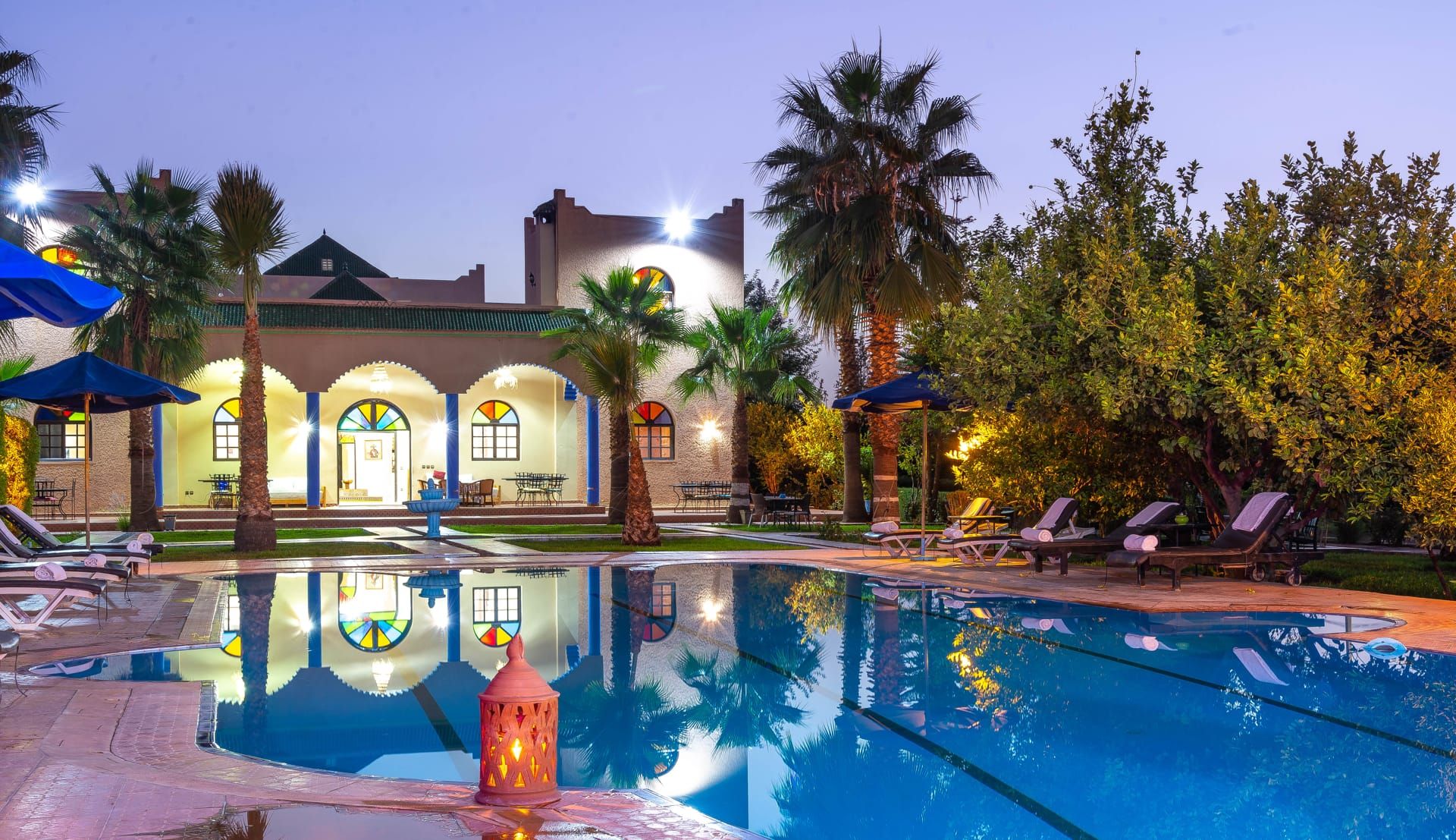 A pool area with lounge chairs, a fountain, and a building with arched windows and a tiled roof.