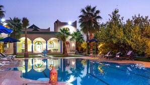 A pool area with lounge chairs, a fountain, and a building with arched windows and a tiled roof. - Riad Qodwa (Sidi Abdallah Ghiat)