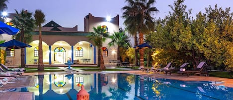 A pool area with lounge chairs, a fountain, and a building with arched windows and a tiled roof.