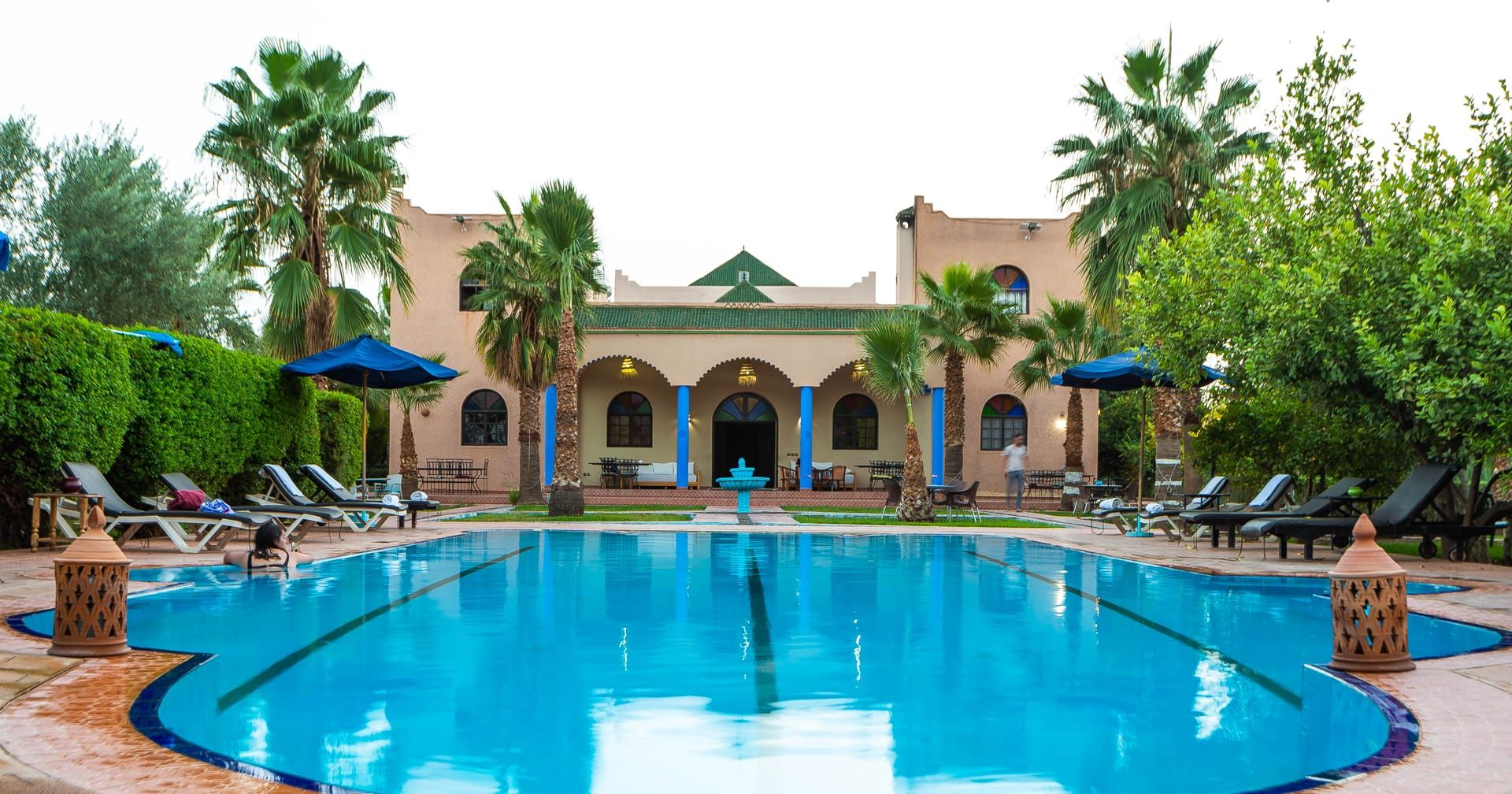 Une piscine entourée de chaises longues et de parasols, bordée de palmiers et d’un bâtiment orné d’arcades.