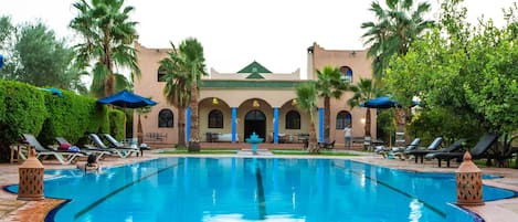 A swimming pool with lounge chairs and umbrellas, surrounded by palm trees and a building with arches.