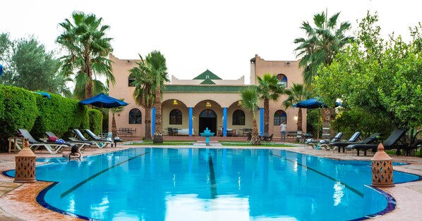 A swimming pool with lounge chairs and umbrellas, surrounded by palm trees and a building with arches. - Riad Qodwa (Sidi Abdallah Ghiat)