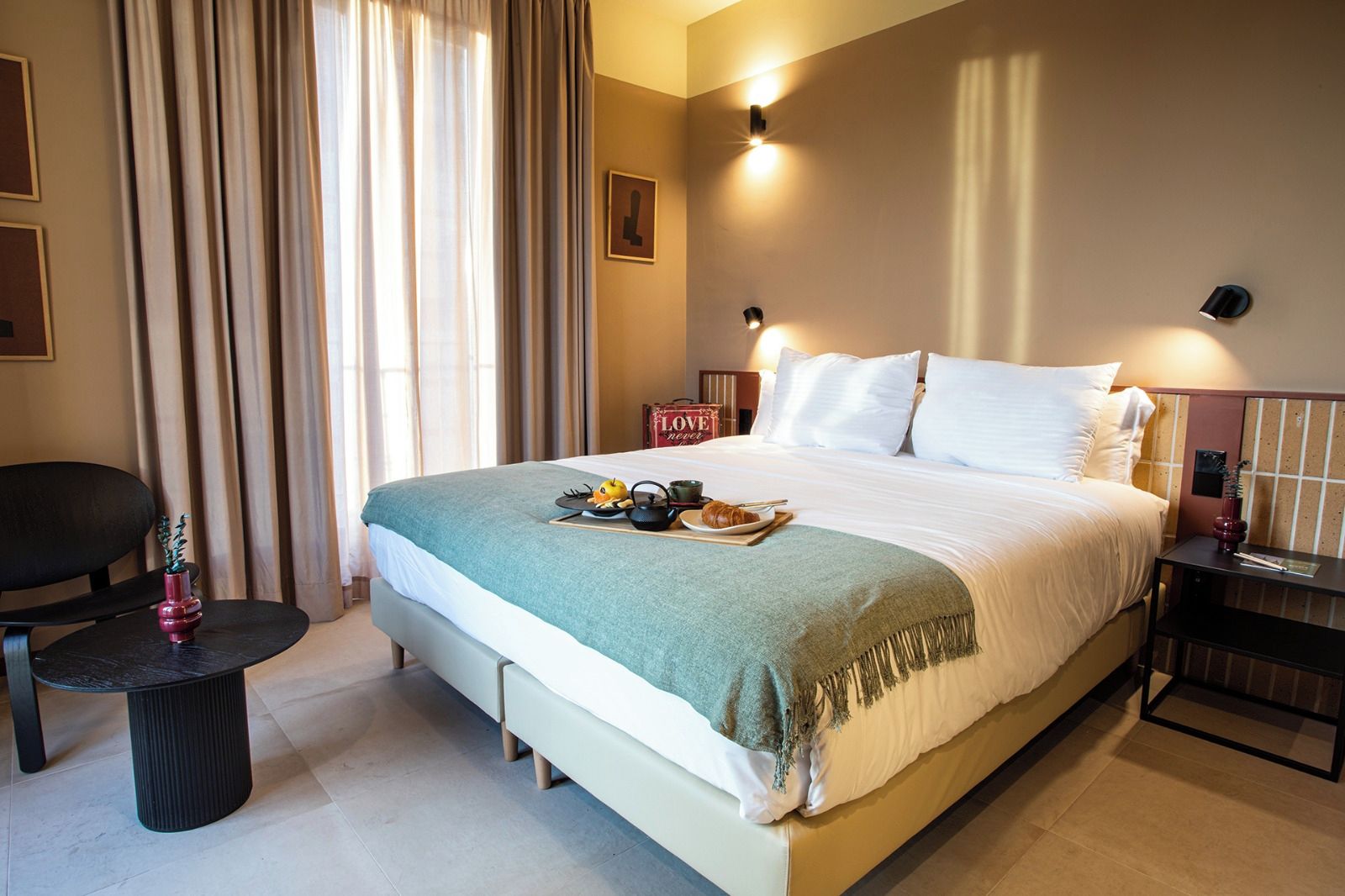Una habitación de hotel con una cama grande, una mesita de noche con lámpara, una silla y una ventana con cortinas.