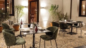 Un área de comedor al aire libre bien iluminada con mesas preparadas para dos, rodeada de plantas en macetas y con sillas tapizadas en verde.