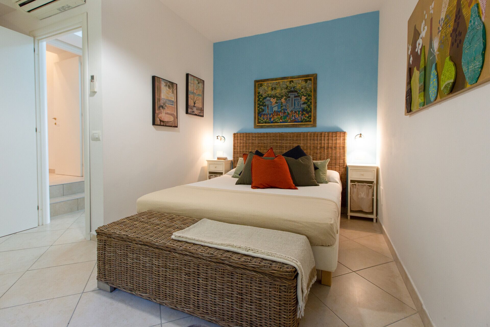 Un dormitorio con una cama, dos mesitas de noche, un banco de mimbre y un cuadro en la pared.