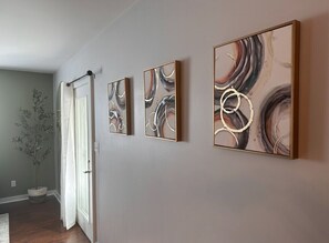 A hallway with three abstract paintings on the wall, a potted plant, and a door with a curtain. - Relax and Roscoe Apartment (Coshocton)