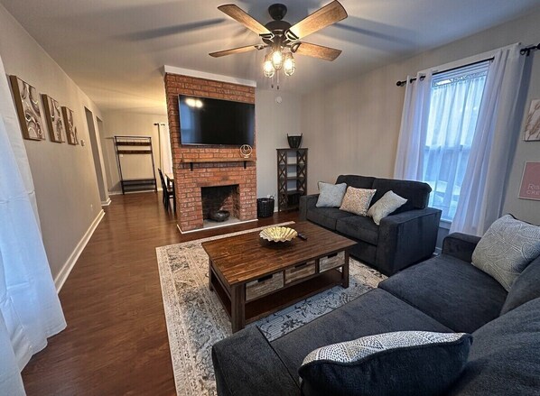 A living room with a brick fireplace, a ceiling fan, and a large flat-screen TV.