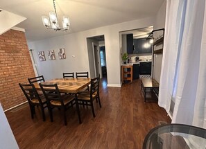 A dining area with a wooden table and chairs, a chandelier, and a kitchen in the background.