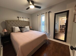 A bedroom with a tufted headboard, a bed with a pink quilt, two bedside lamps, a ceiling fan, a large mirror, and a view of a staircase and outdoor area.
