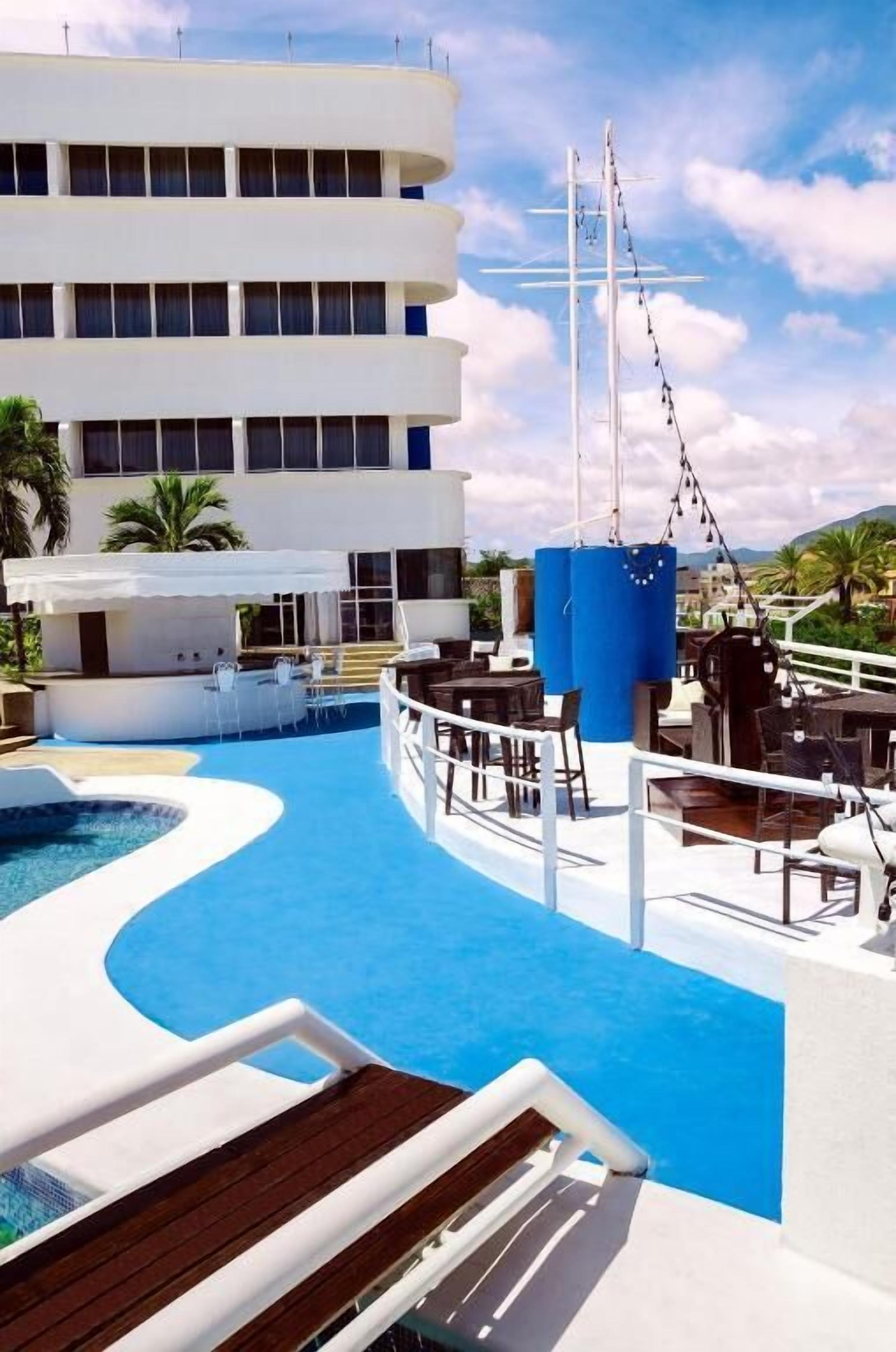 A pool area with a white building in the background, outdoor seating, and a clear sky.