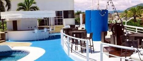 A pool area with a white building in the background, outdoor seating, and a clear sky.