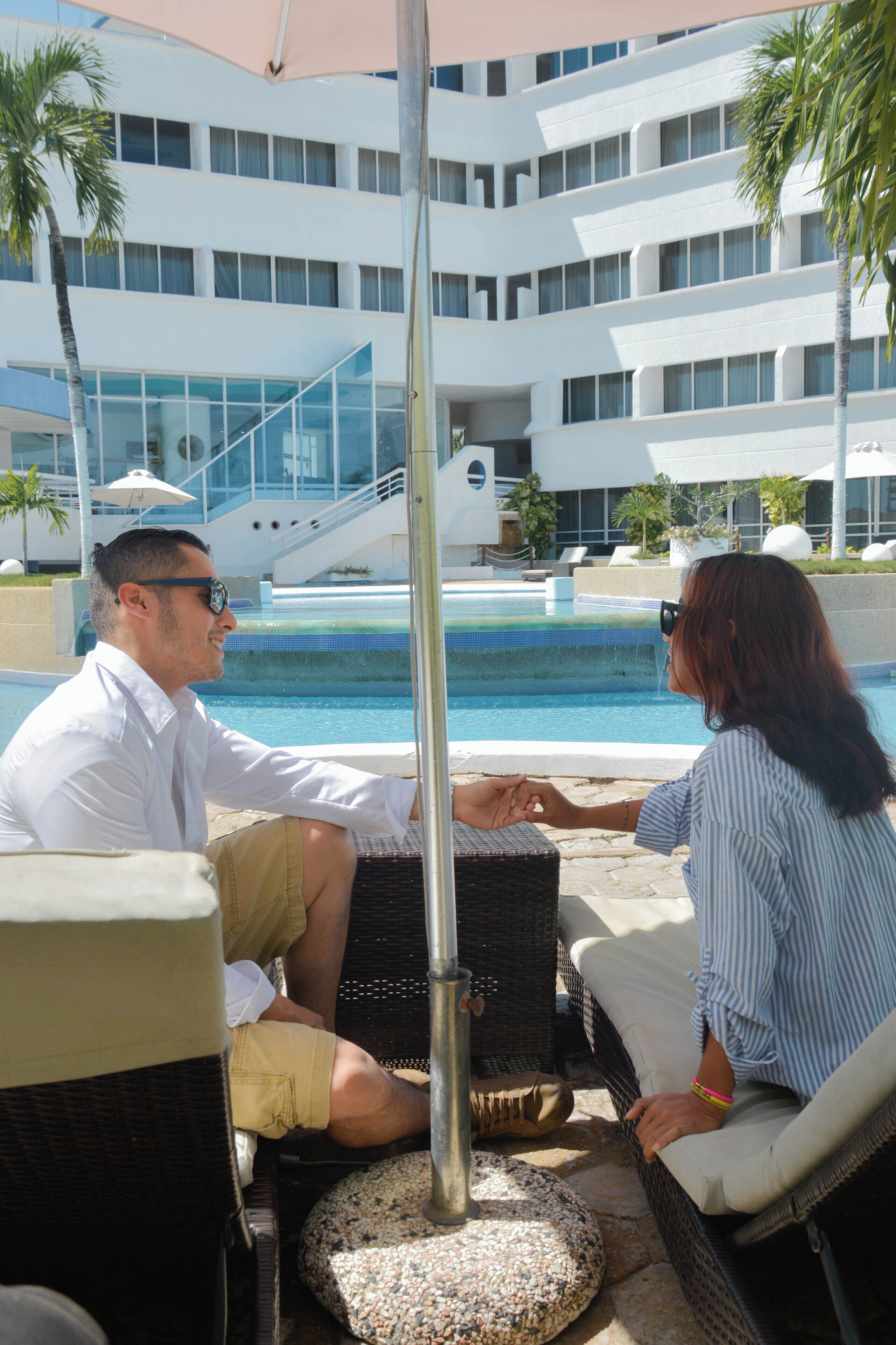 Un couple est assis à une table au bord de la piscine, avec une vue sur un bâtiment moderne et une piscine en arrière-plan.
