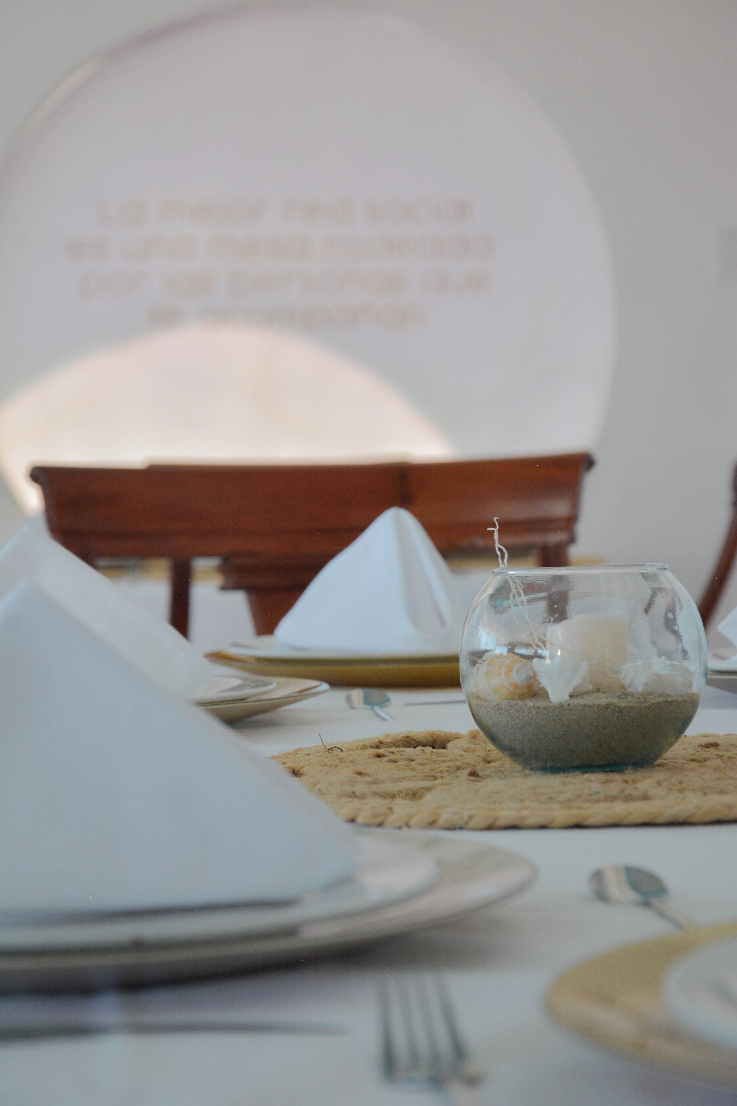 A dining table set with plates, cutlery, and a centerpiece of a glass bowl with sand and shells.