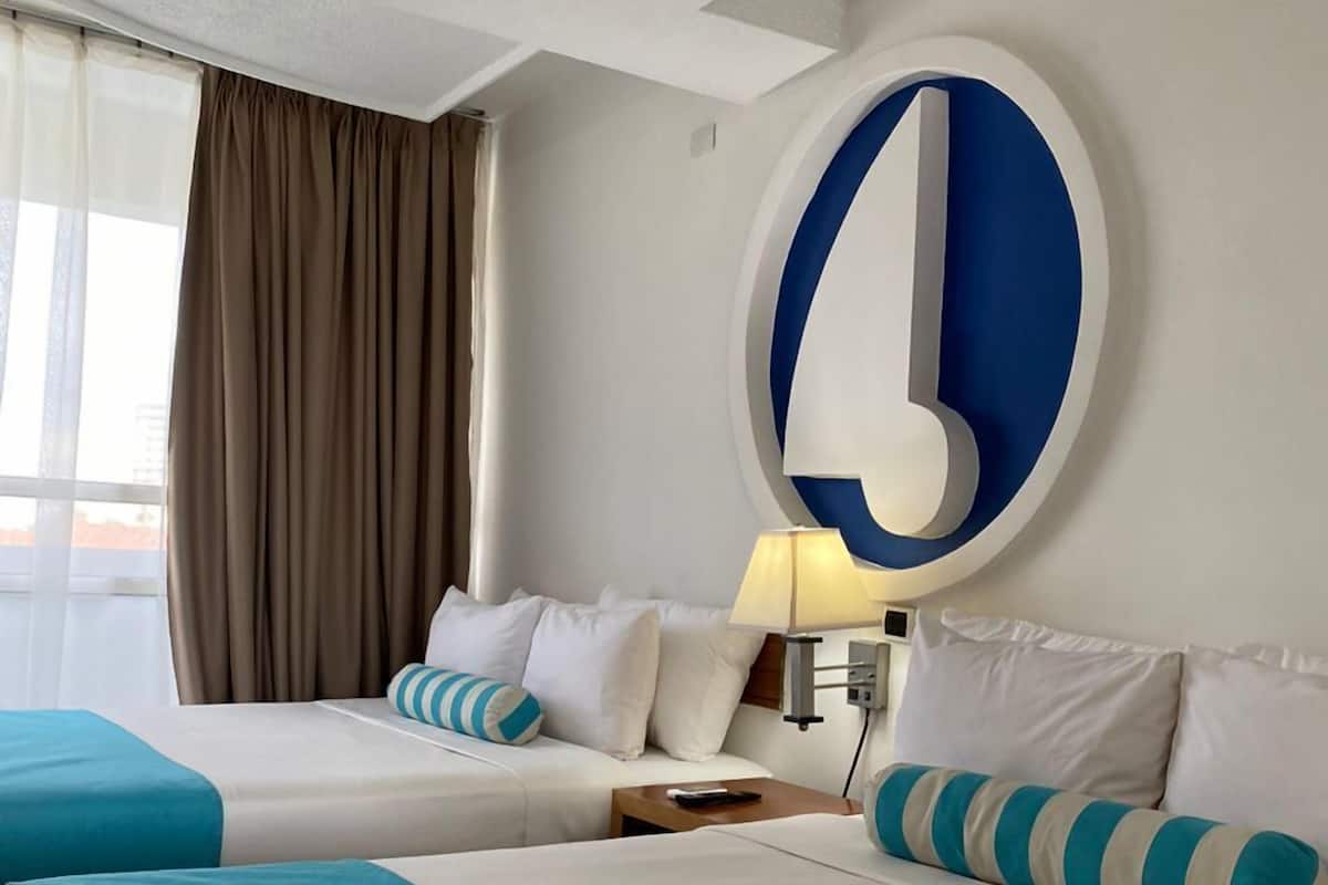 Habitación de hotel con dos camas, un reloj de pared grande, una lámpara y una ventana con cortinas.