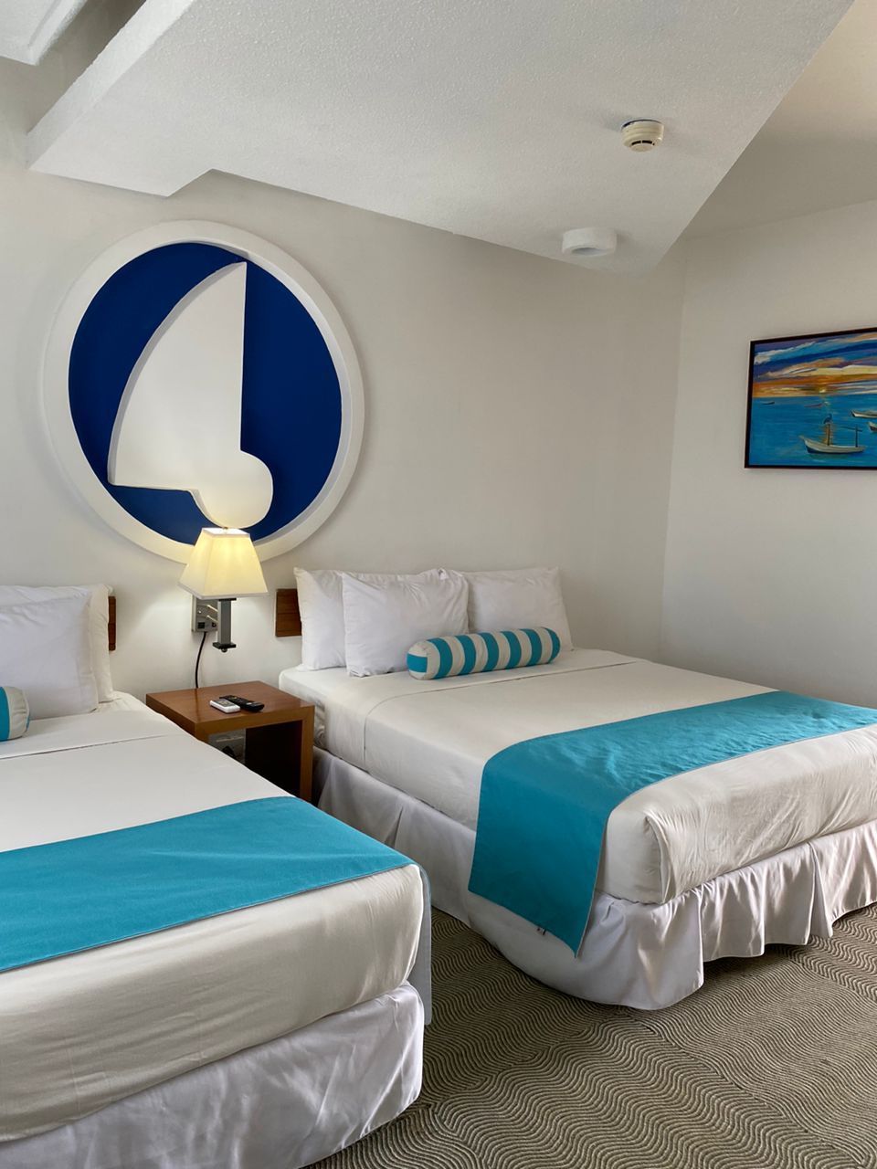 A hotel room with two beds, a large wall clock, a painting, and a ceiling light.