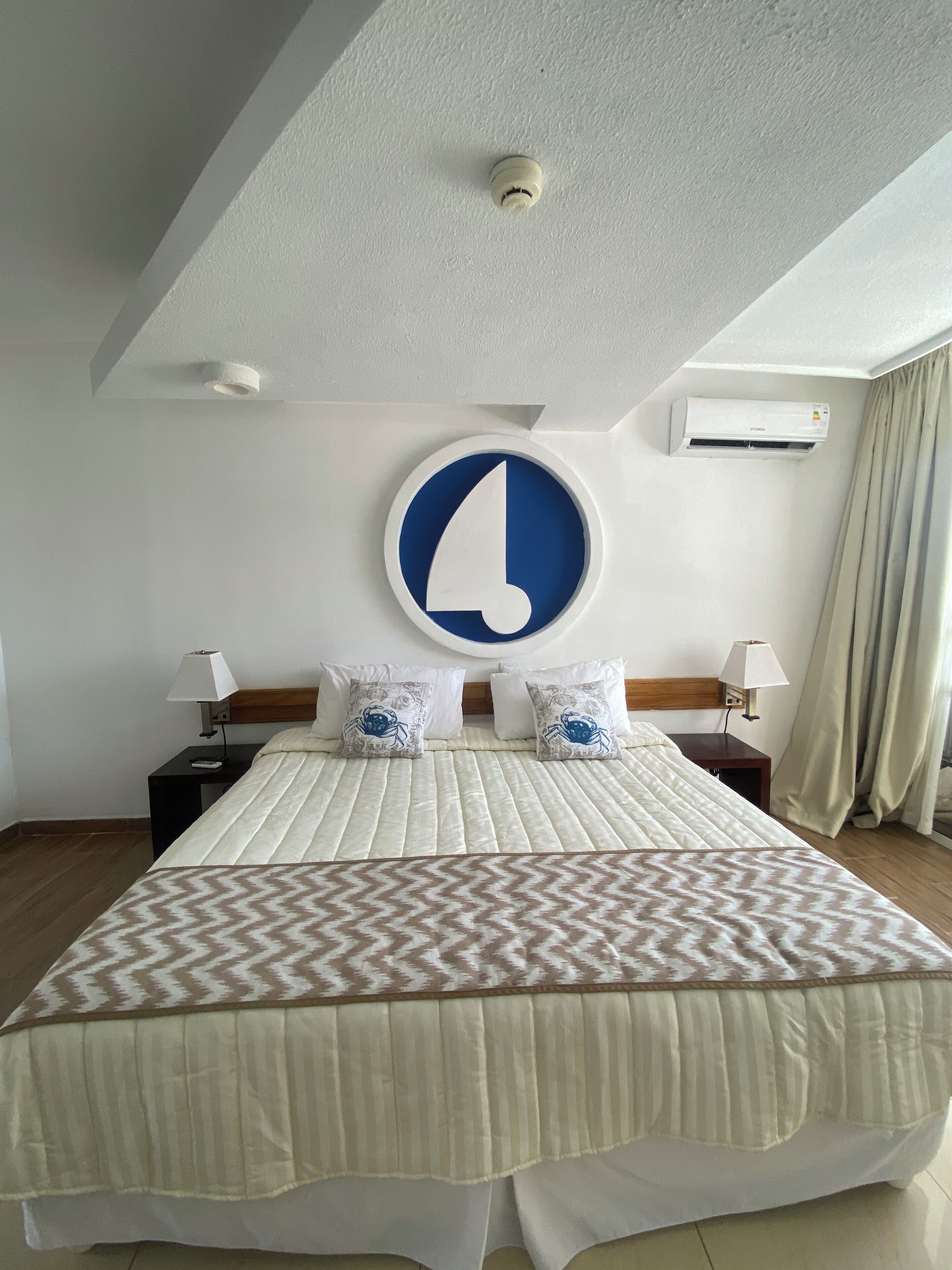 A hotel room with a large bed, a round wall clock, an air conditioning unit, and a nightstand with a lamp.