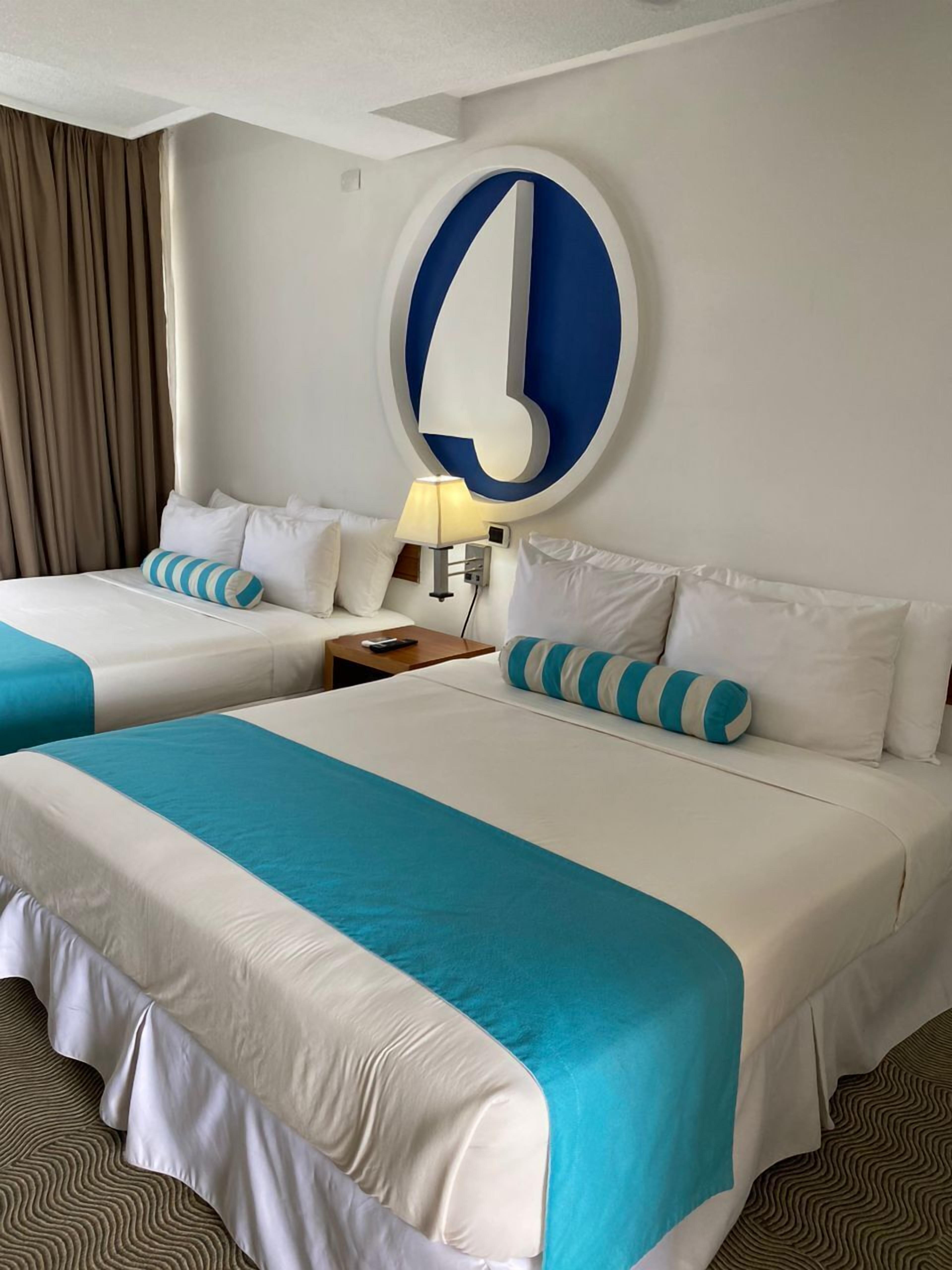 Ein Hotelzimmer mit zwei Betten, jeweils mit weißer Bettwäsche und blauen Akzenten. Darüber hängt eine große, blau-weiße Wanddekoration.