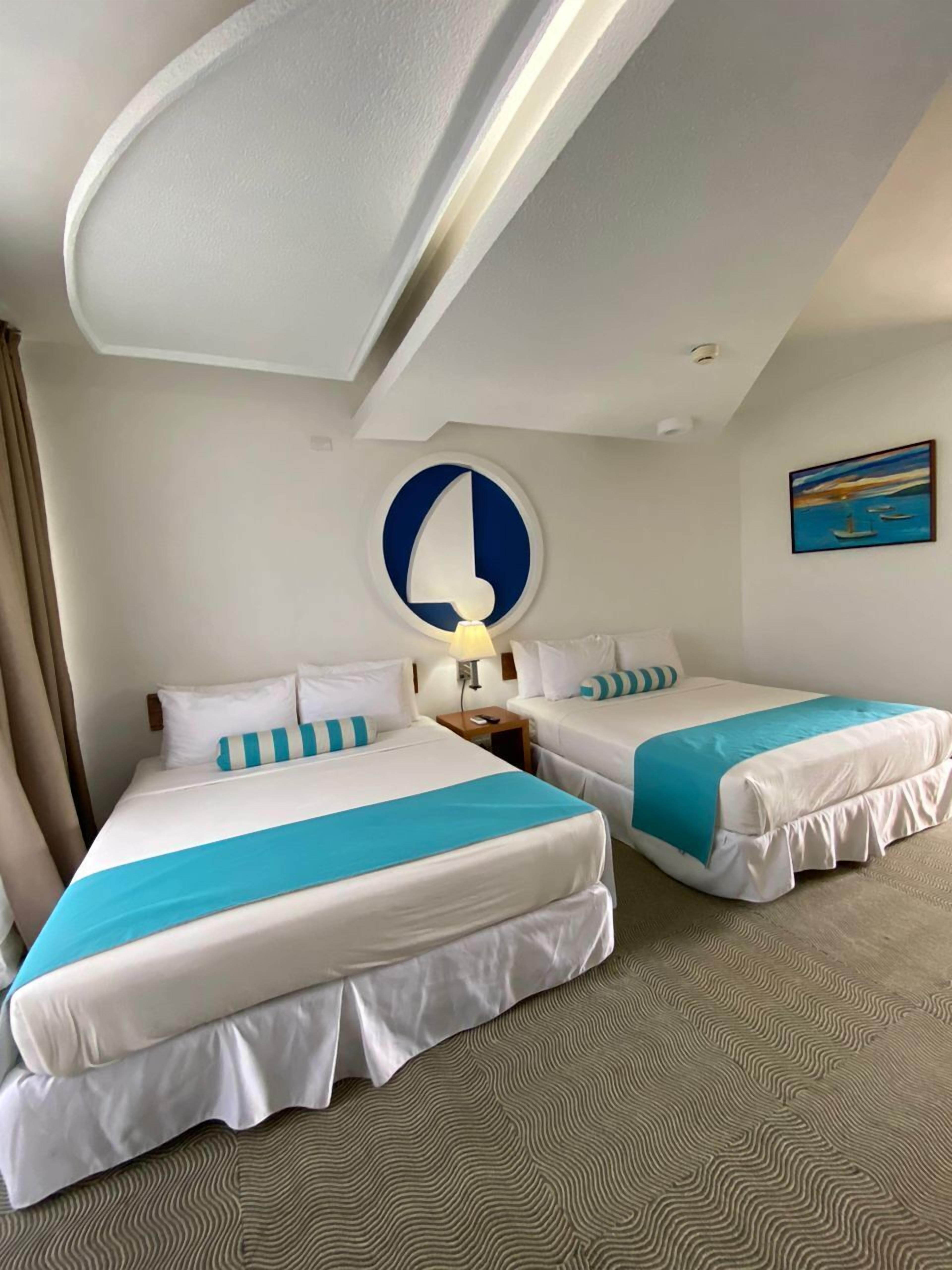 A hotel room with two beds, a ceiling light, a wall clock, and a painting.
