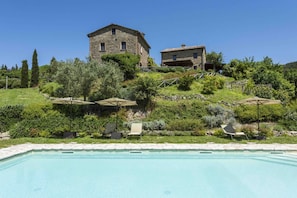Uma área de piscina com espreguiçadeiras e guarda-sóis, um edifício de pedra e vegetação exuberante.