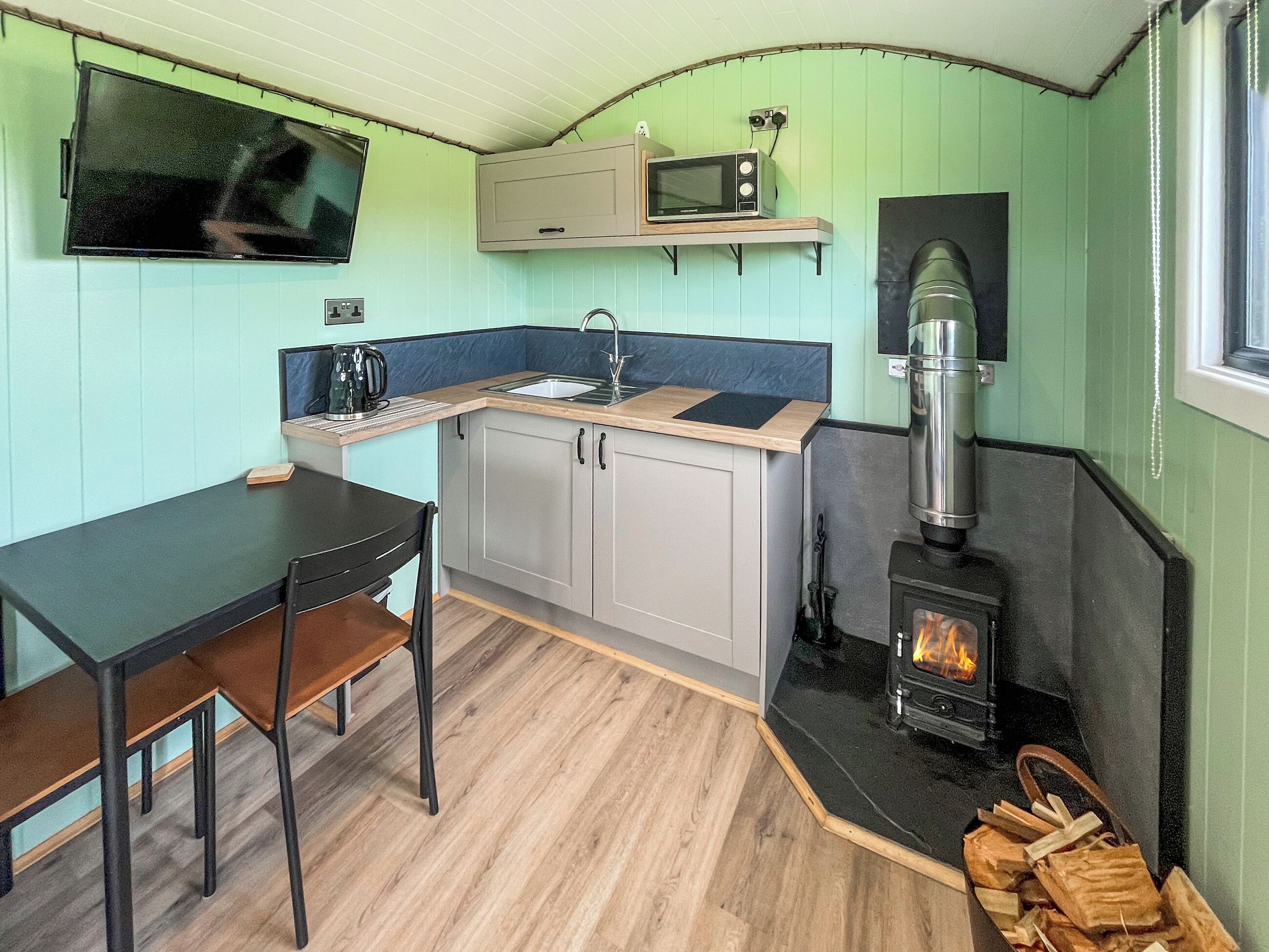 A compact kitchen with a wood-burning stove, a microwave, and a TV mounted on the wall.