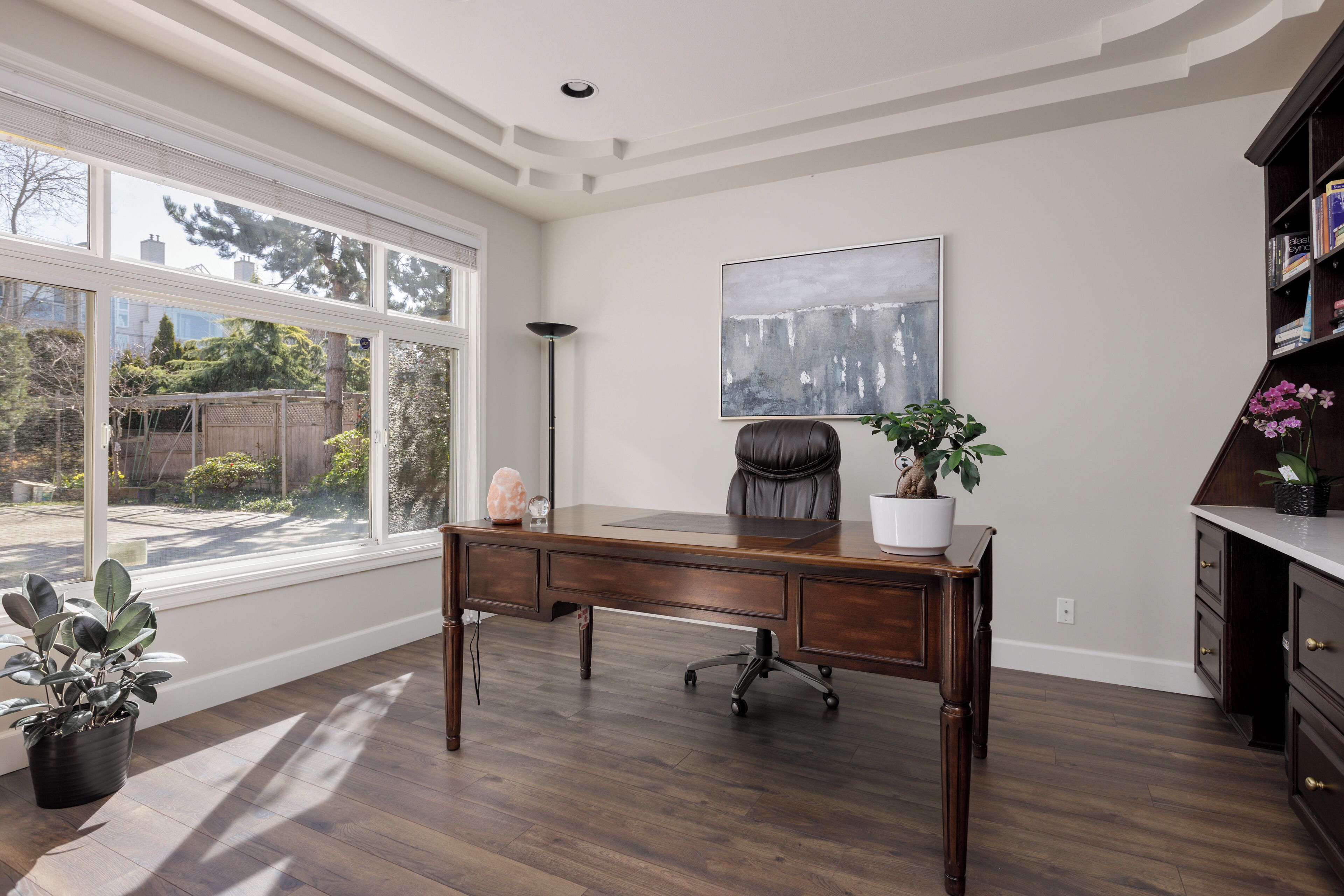 A spacious room with a large window, a wooden desk, a chair, a potted plant, and a bookshelf.