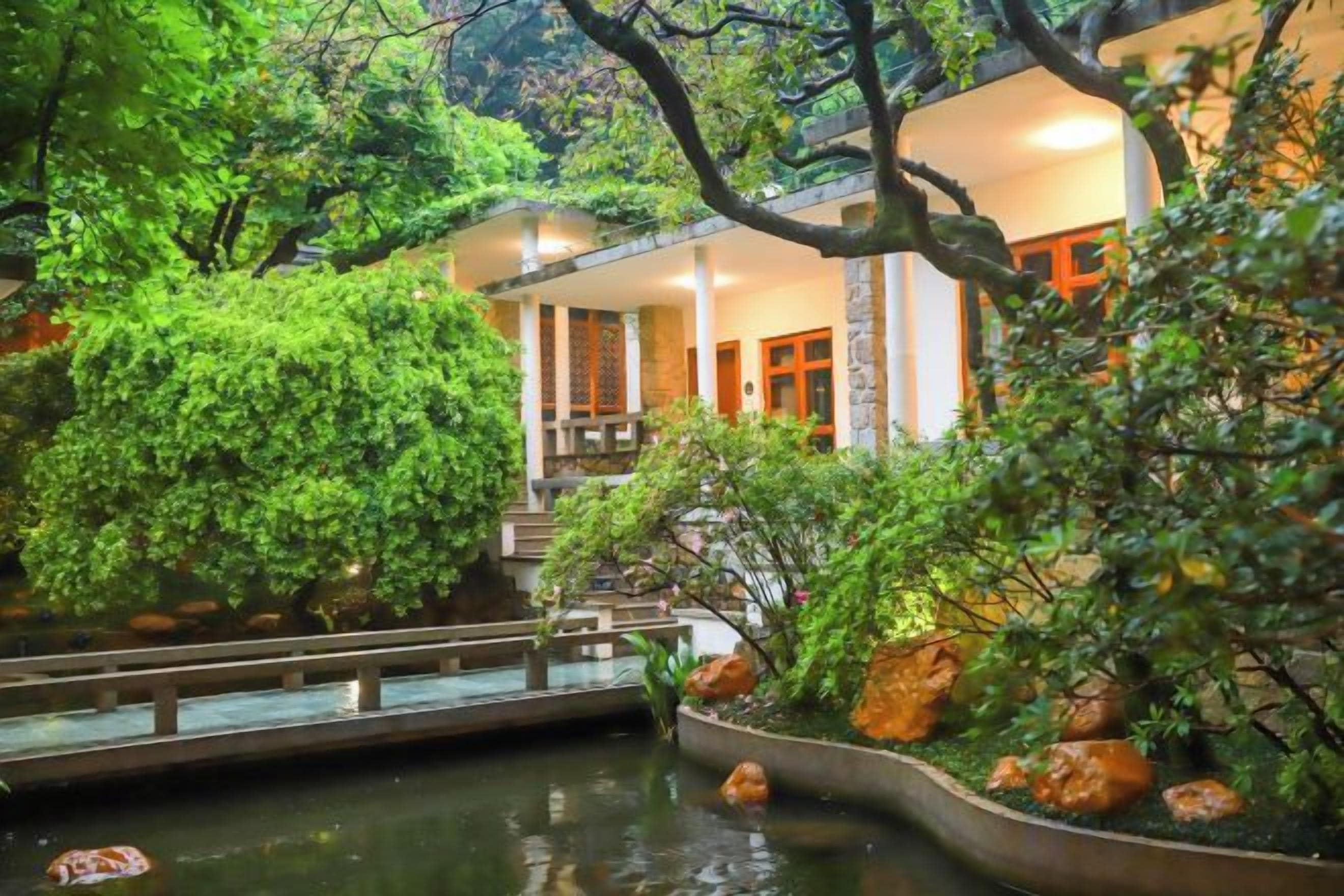 Un edificio de estilo japonés tradicional con un estanque de carpas, rodeado de exuberante vegetación y un sendero de piedra.