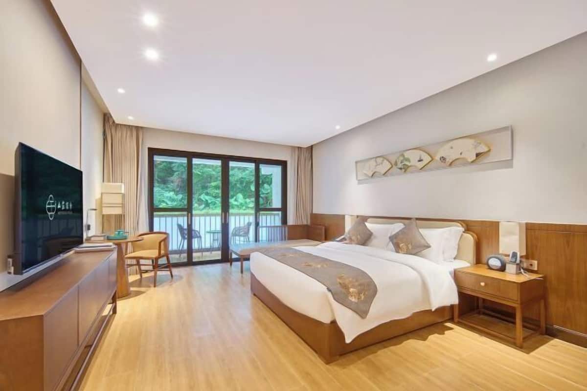 Une chambre d’hôtel moderne avec un grand lit, un bureau doté d’un téléviseur à écran plat, une chaise et une vue sur la verdure par la fenêtre.